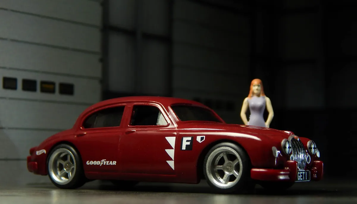 A red toy car with 'GOOD YEAR' and 'F' decals is shown in front of a garage door, with a small figurine of a woman in a…