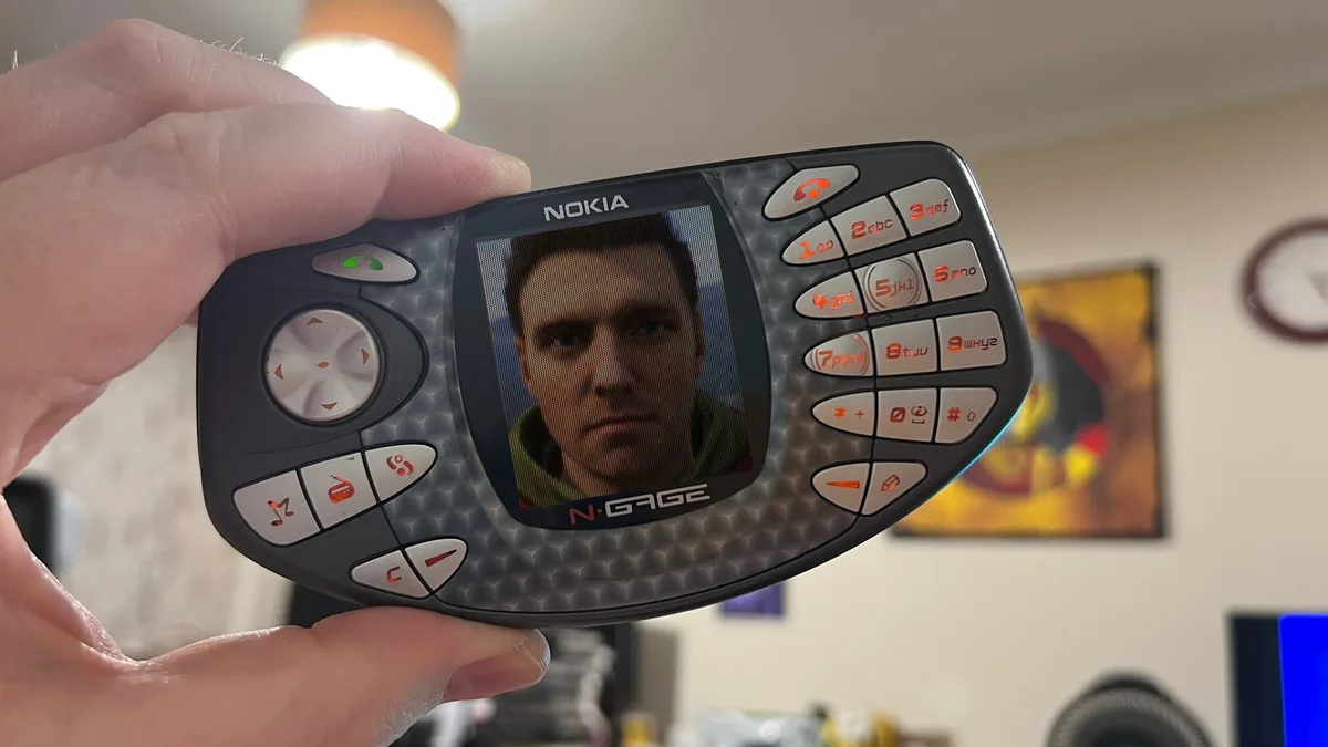 A hand holds a black Nokia N-Gage phone, its screen displaying a close-up of a man's face with a serious expression. The…