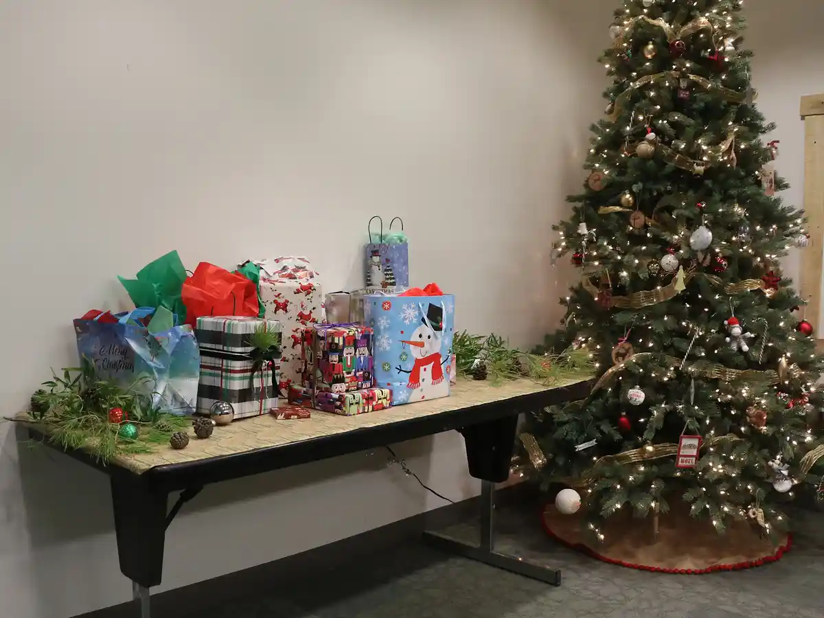 A table next to a decorated Christmas tree is piled high with wrapped gifts and festive bags. The tree is adorned with…