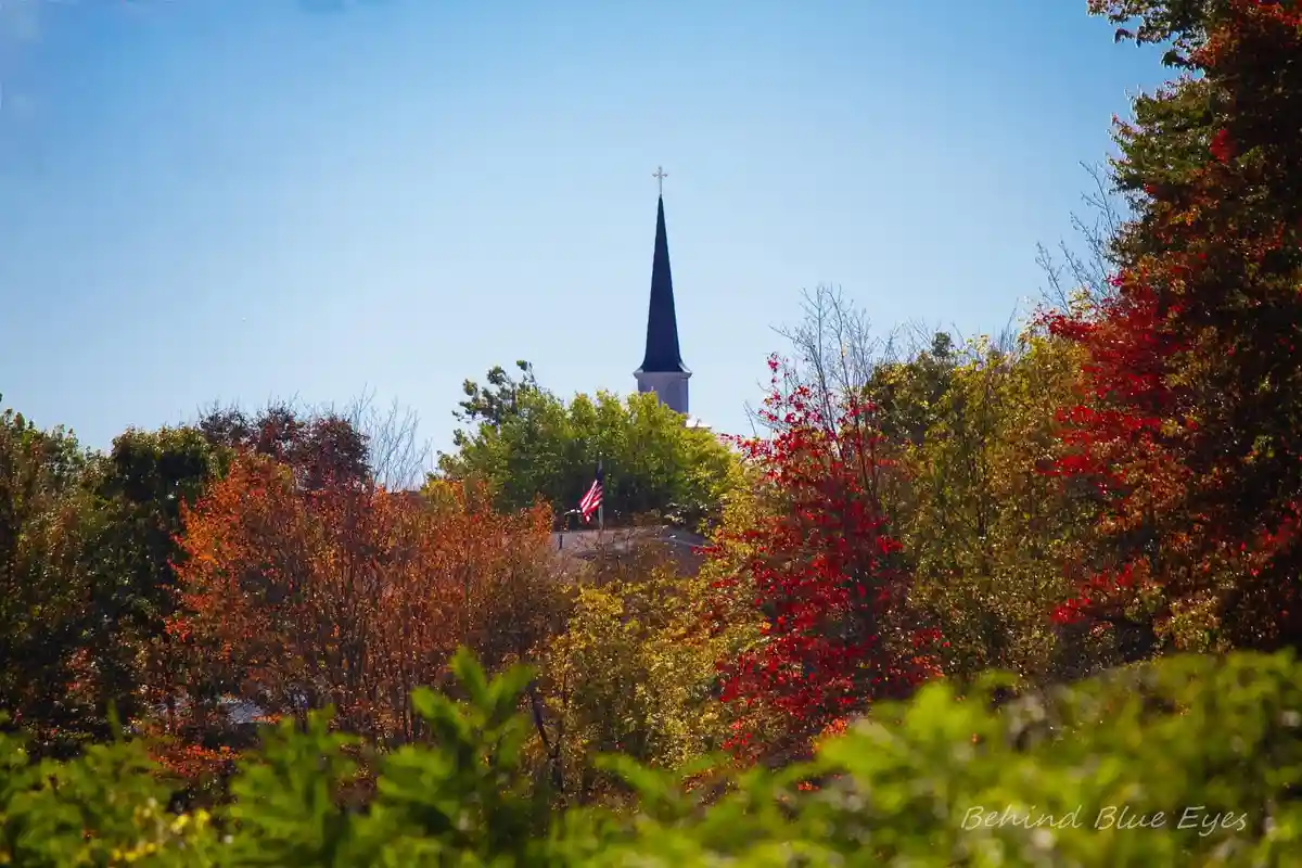 A scenic autumn photo shows a church steeple with a cross peeking above colorful trees with red, orange, and green leaves…
