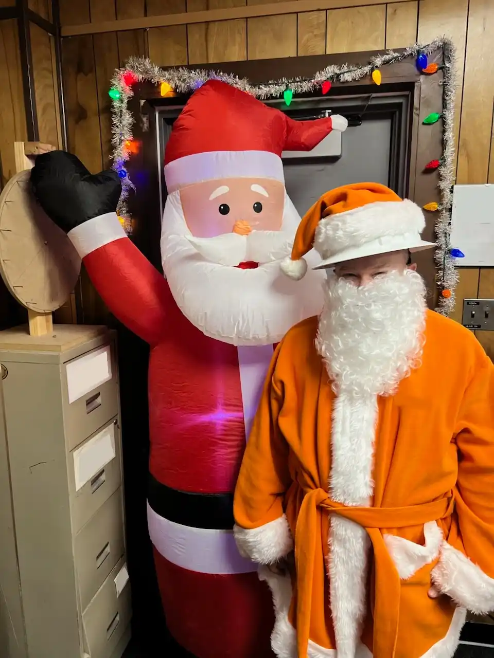 A person in an orange Santa costume with a white beard and hat stands beside a large inflatable red Santa Claus. The person…