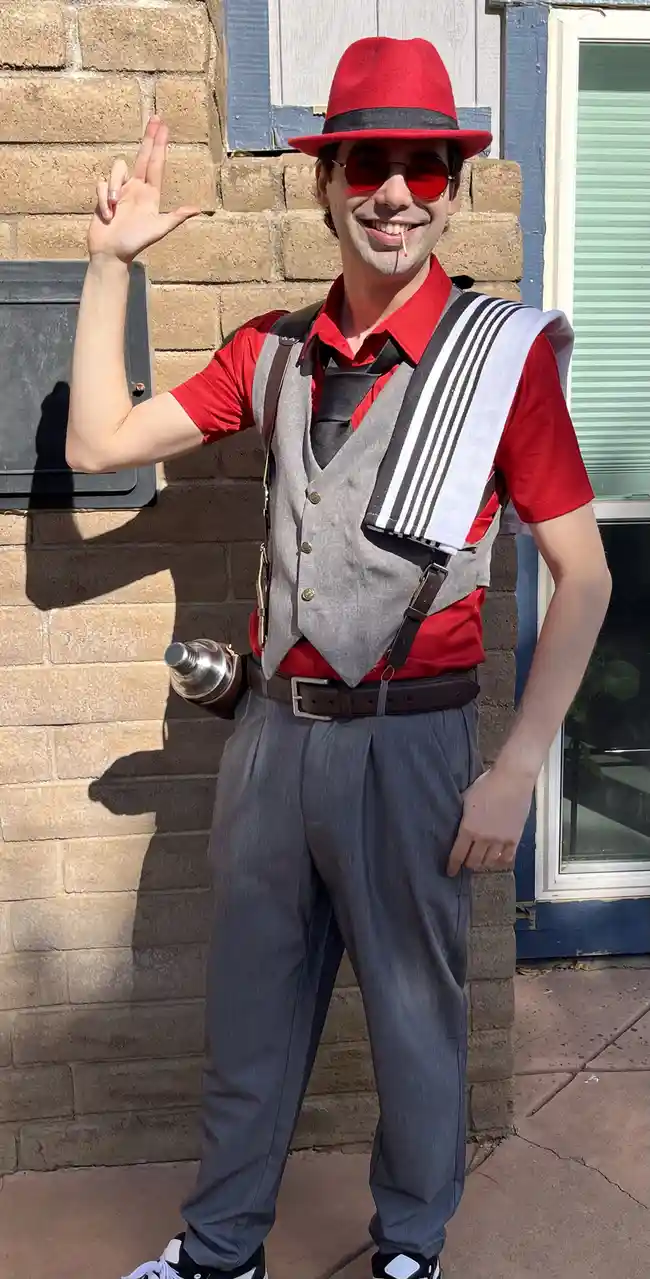 A man in a red fedora, red sunglasses, and a red shirt under a grey vest poses outdoors against a brick wall. He holds up…