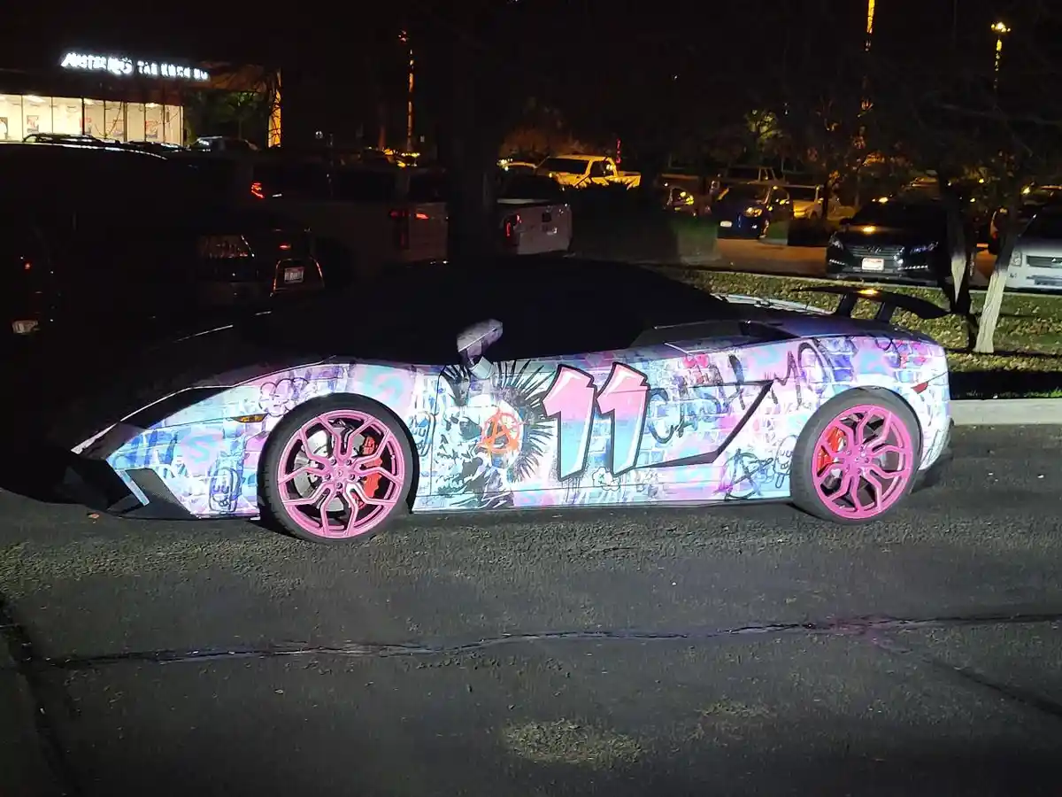 A Lamborghini convertible parked at night is covered in colorful graffiti-style artwork, featuring the text '11 cash mo' in…