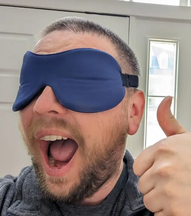 A man with a beard and short hair wears a blue sleep mask over his eyes while giving a thumbs-up gesture with his right…