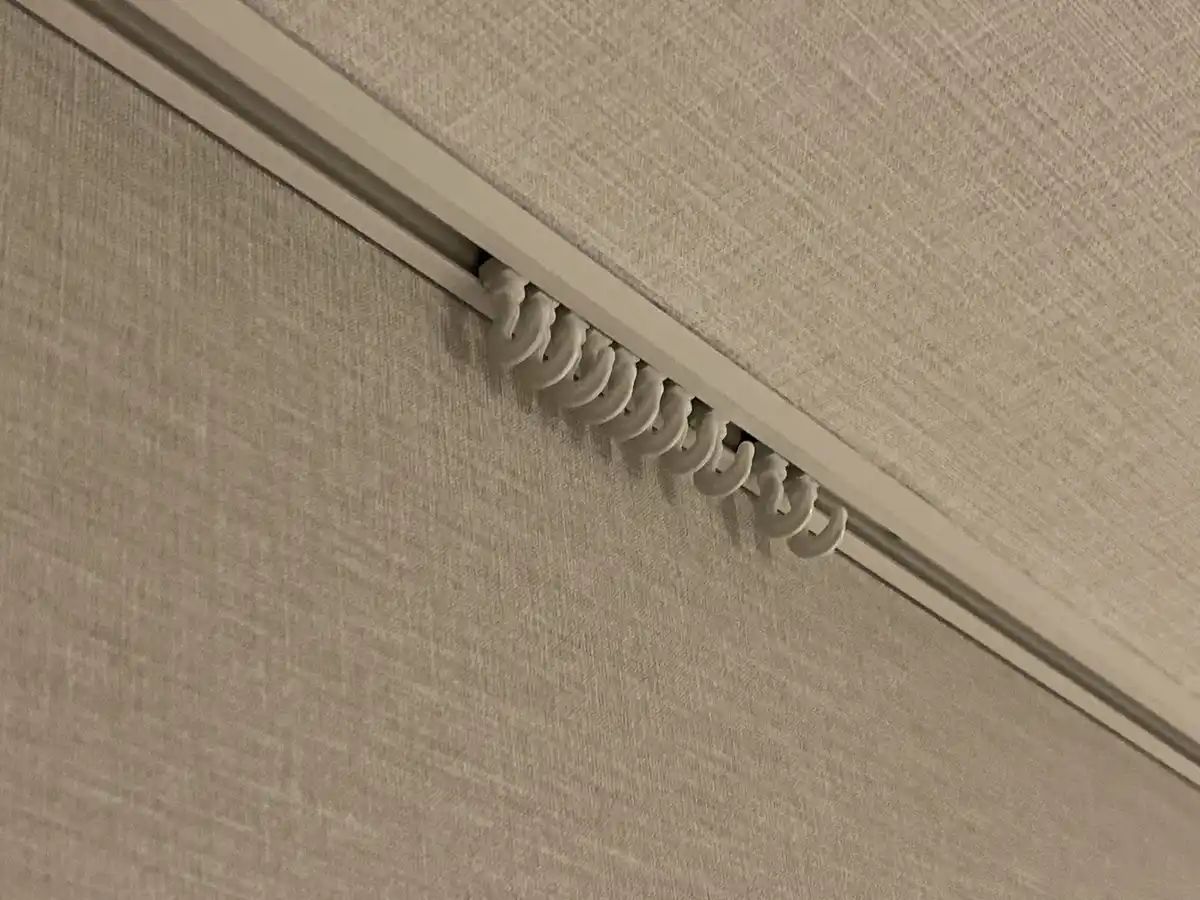 A close-up photo shows a row of white, U-shaped curtain hooks protruding from a track embedded in a beige textured wall or…