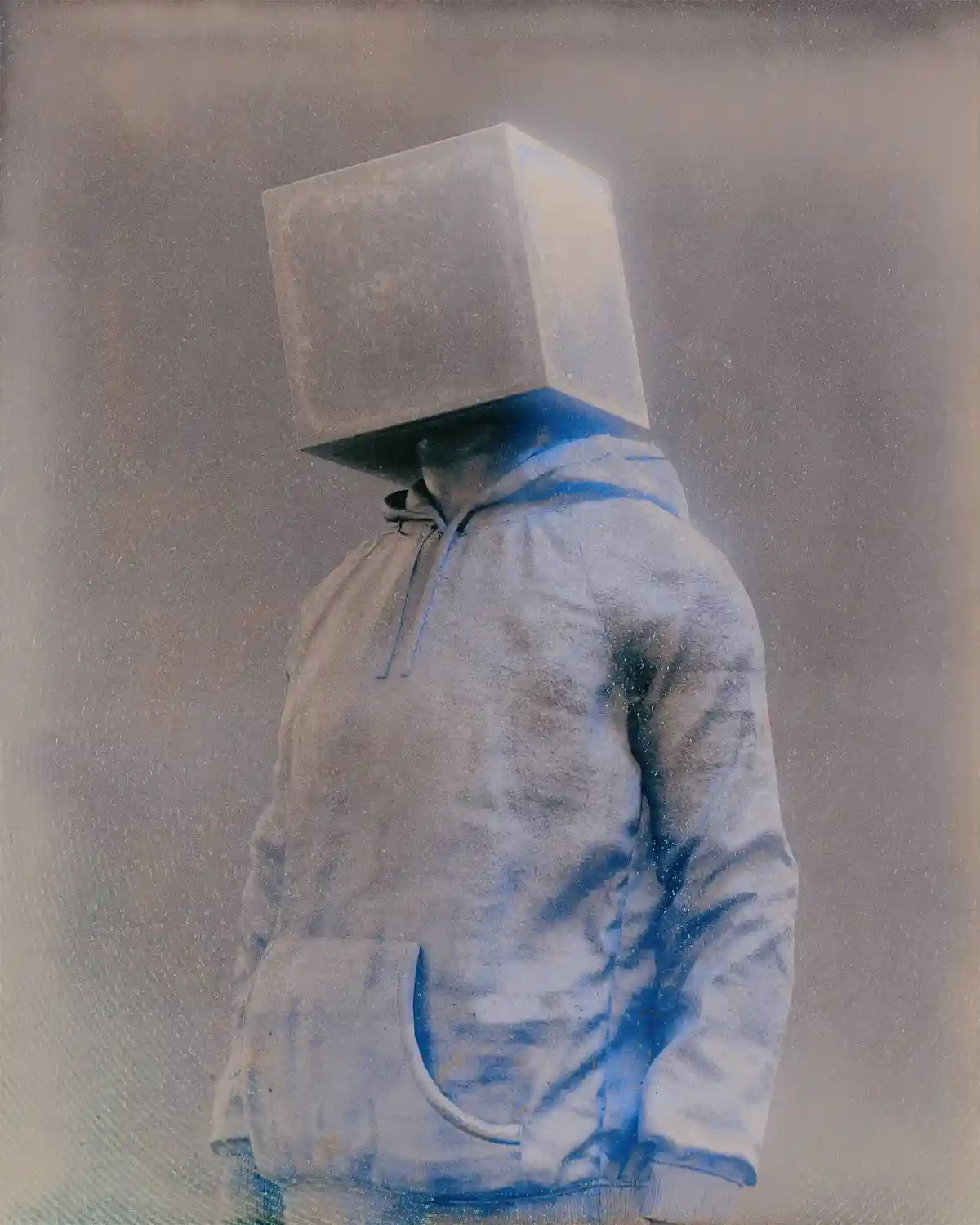 A person wearing a blue hoodie has a large, plain gray cube for a head, standing against a muted, grainy background. The…