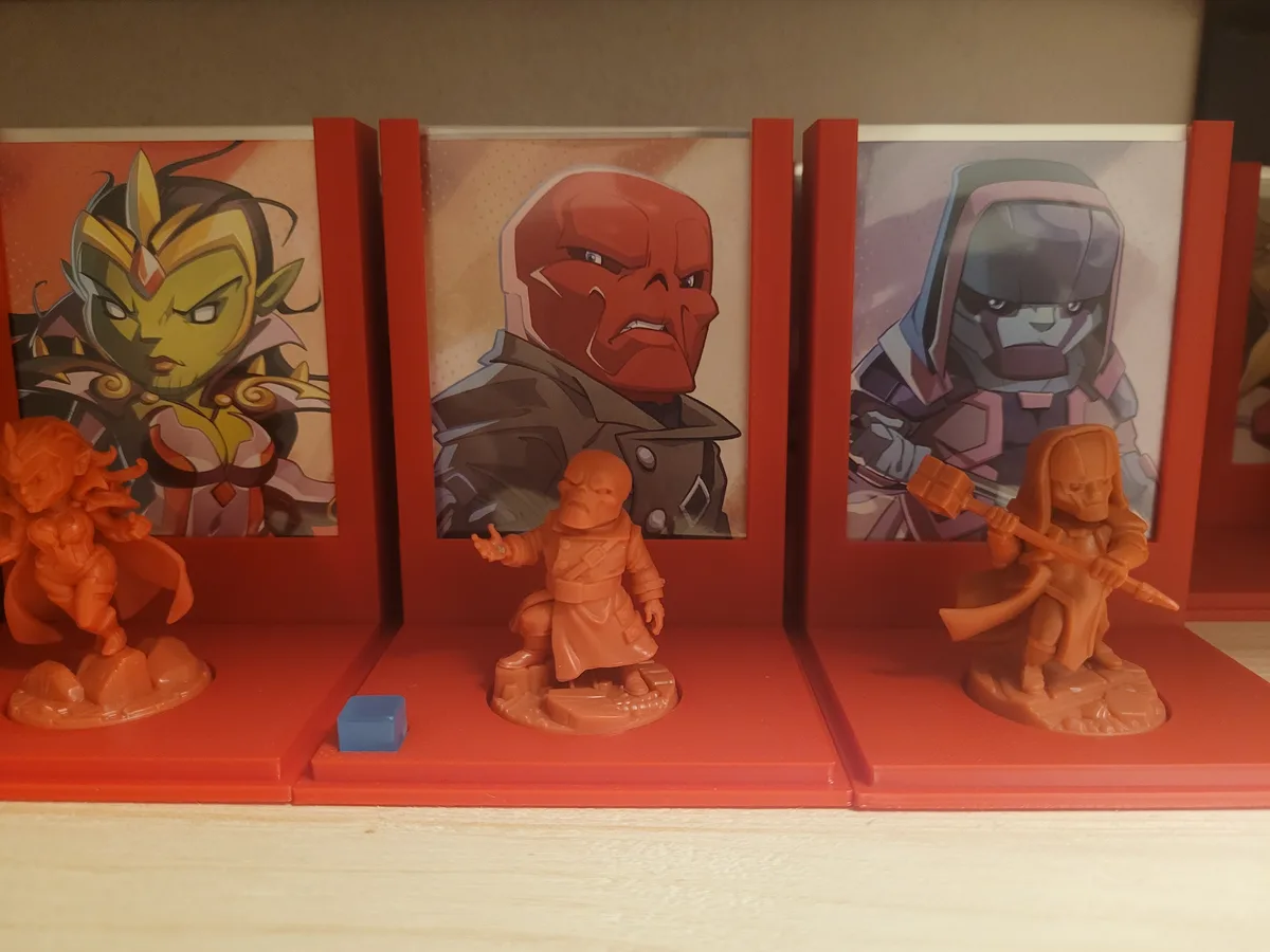 Three orange plastic miniatures stand before red-framed portraits of comic book characters. The middle miniature has a blue…