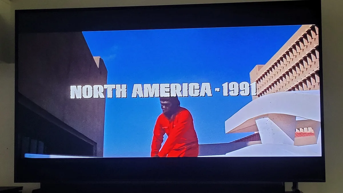 A television screen displays a scene from a video game with a character in a red outfit standing between stylized buildings…