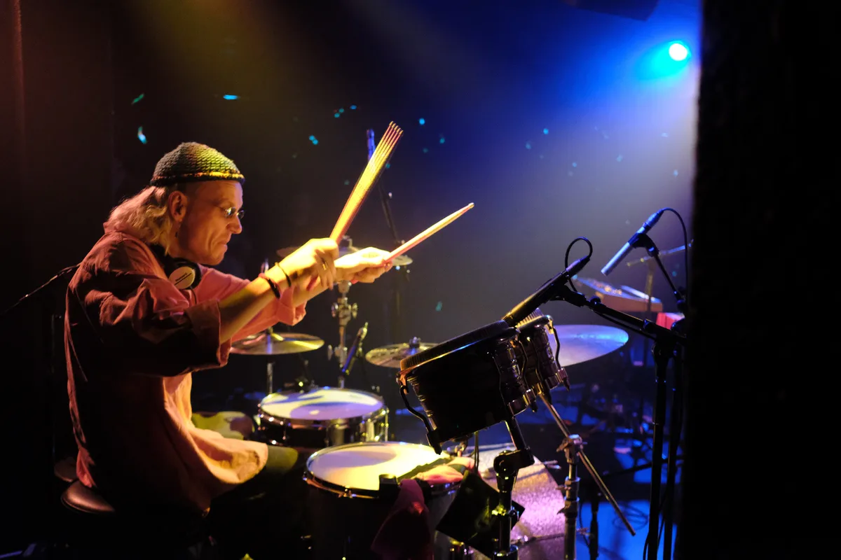 A drummer with long hair, glasses, and a knit cap sits at a drum kit under stage lights, holding drumsticks in a relaxed…