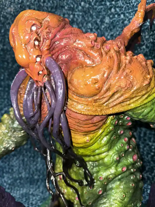 A close-up of a grotesque, multi-colored monster figurine with an orange, fleshy head covered in white eyes, purple…