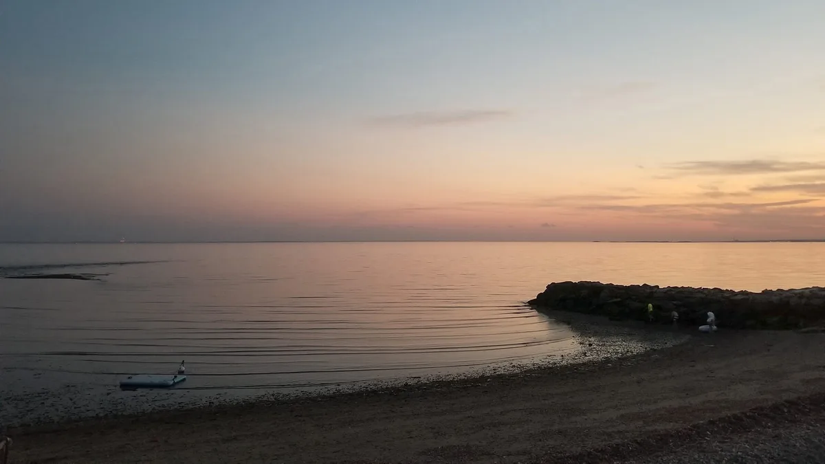 A serene beach at sunset with calm water reflecting orange and pink hues, a rocky jetty on the right, and a small inflatable…
