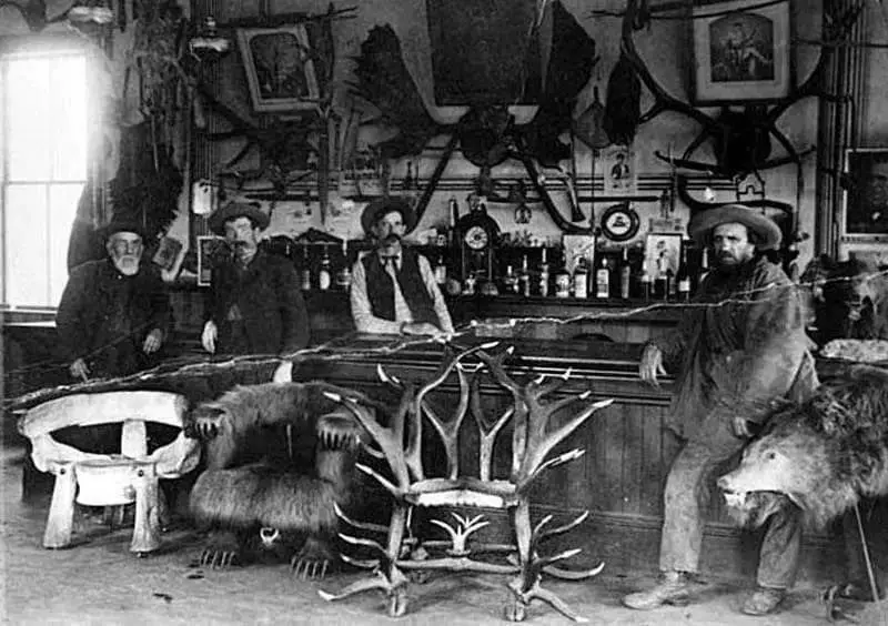 A vintage black-and-white photo shows four men in a rustic bar decorated with mounted animal heads and antlers. Two of the…
