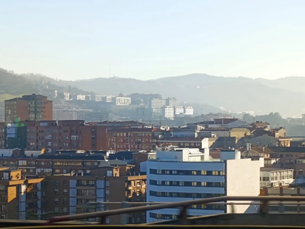 A cityscape photo showing a mix of residential buildings with hills in the background under a hazy sky, overlaid with the…