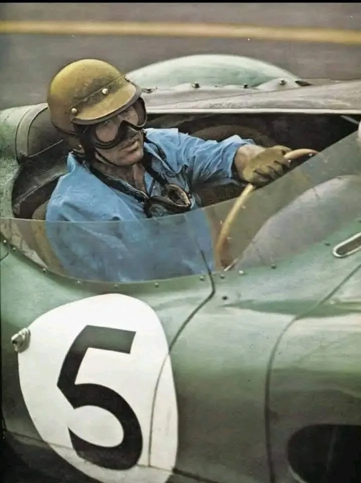 A vintage photograph shows a male race car driver in a light blue shirt, yellow helmet, and goggles, gripping the steering…
