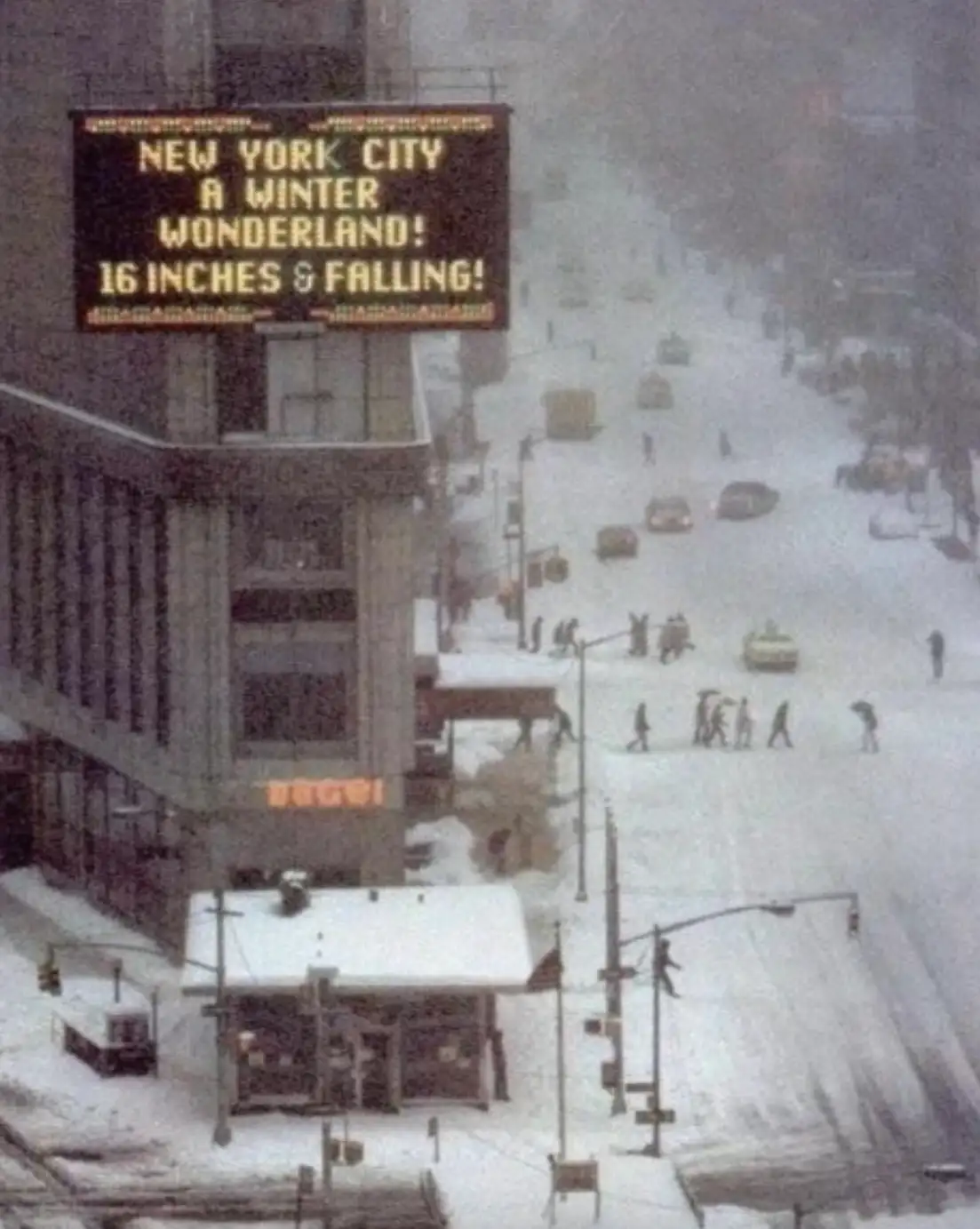 A snowy, chaotic street scene in New York City with people and cars barely visible under heavy snowfall. A large electronic…