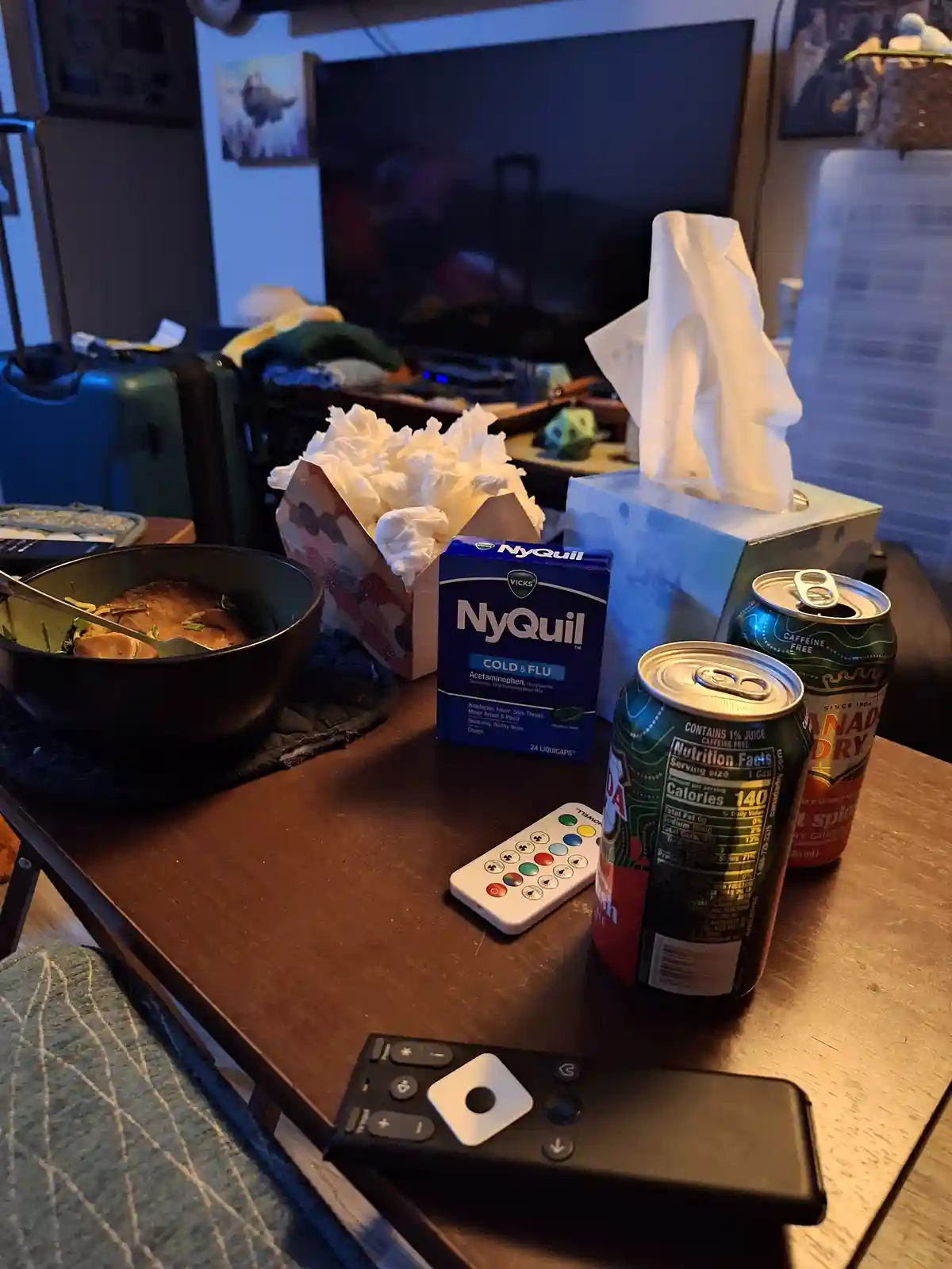A cluttered living room table holds a bowl of food, two cans of Canada Dry ginger ale, a box of Vicks NyQuil cold & flu…
