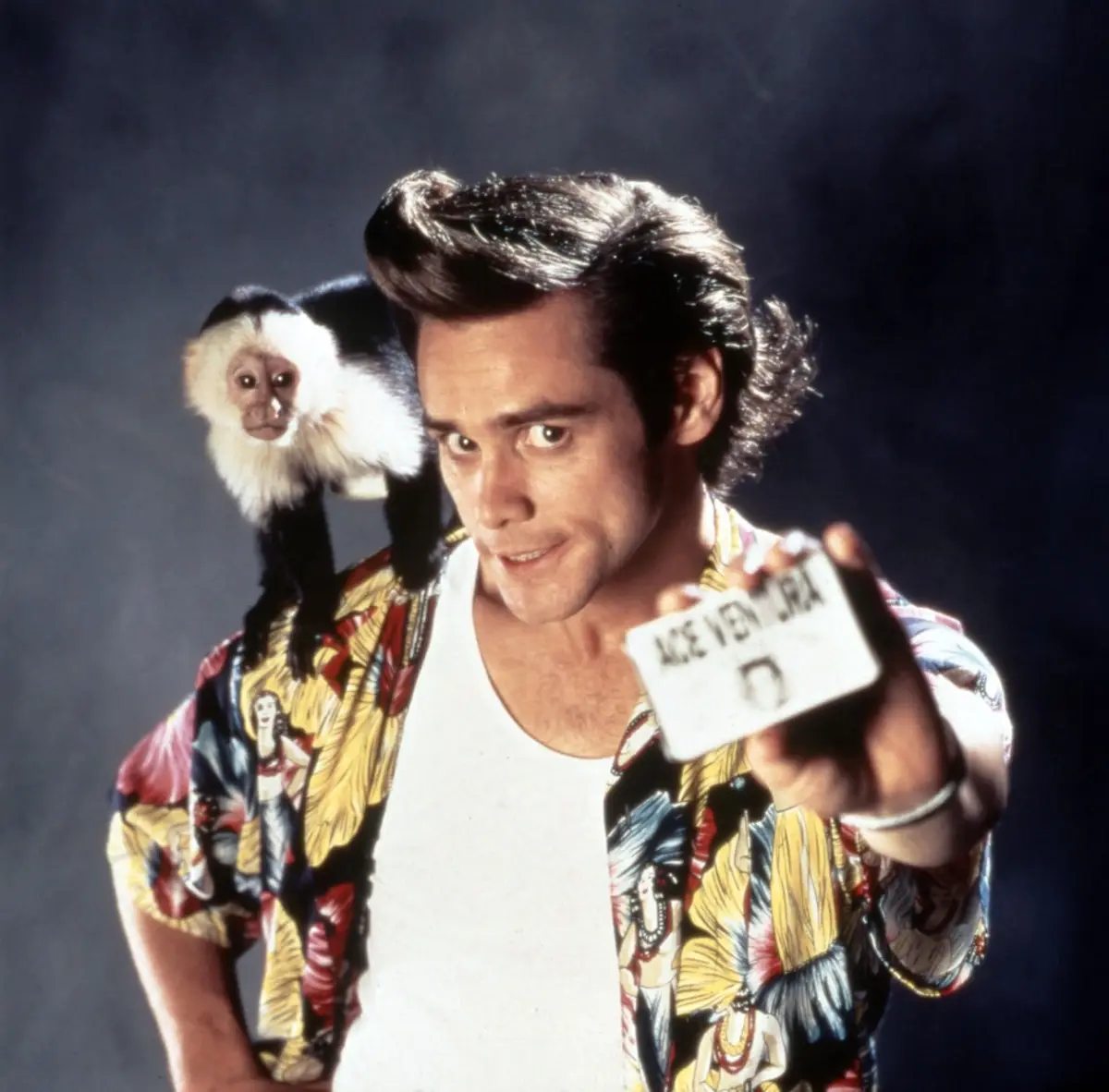 A man with a distinctive pompadour hairstyle and a colorful patterned shirt holds up a card that reads 'ACE VENTURA' towards…