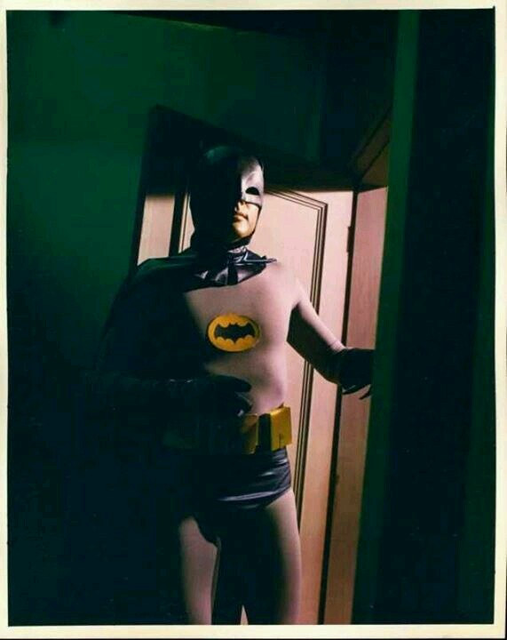 A person in a vintage Batman costume stands in a doorway, the costume missing its chest plate and revealing their bare…