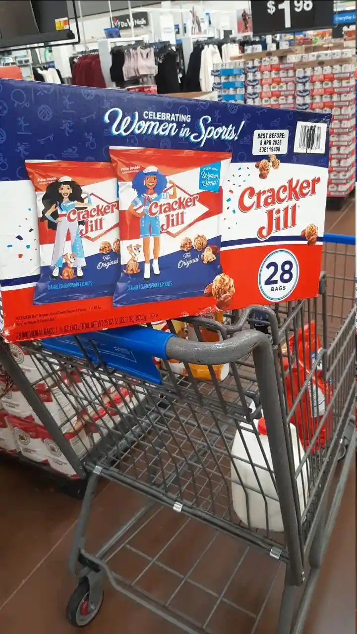 A shopping cart in a store holds a large box of Cracker Jill snack bags, which features illustrations of two women in…