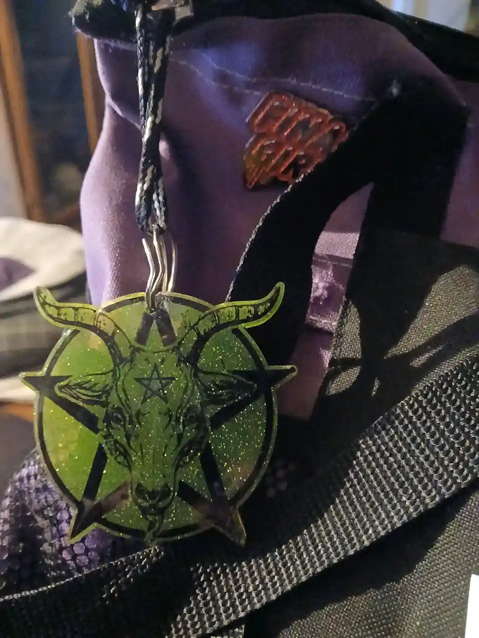 A close-up photo shows a green glittery pentagram pendant with a goat's head and horns hanging from a black and white…