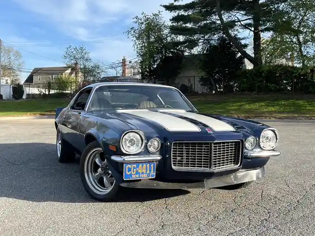 A dark blue classic American muscle car with white racing stripes is parked on an asphalt road, facing forward. The car has…