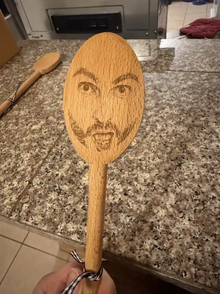 A hand holds a wooden spoon with a crudely burned face etched onto its flat head, showing eyes, eyebrows, a mustache, and an…