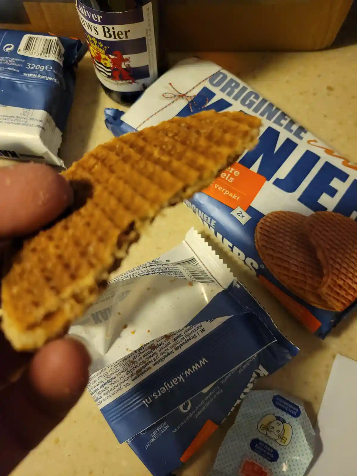A hand holds a partially eaten, thin, grid-patterned waffle cookie with a chocolate filling, above its blue and white…