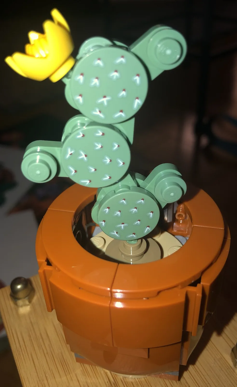 A close-up of a LEGO cactus model with a yellow LEGO crown on top, sitting in an orange pot. The cactus is made of green…
