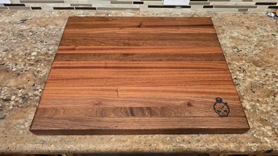 A large, polished wooden cutting board with a distinctive skull-shaped stopwatch logo engraved in the bottom right corner…