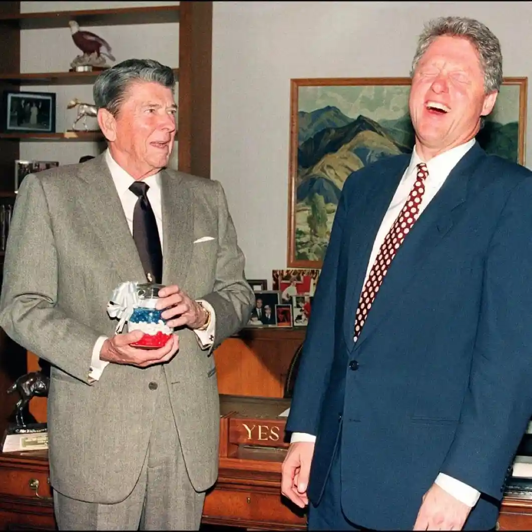 Former U.S. President Ronald Reagan holds a small American flag-themed jar while standing next to former President Bill…