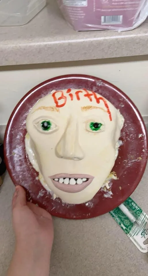 A hand holds a red plate with a partially eaten cake shaped like a human face, featuring green candy eyes, a pink mouth with…