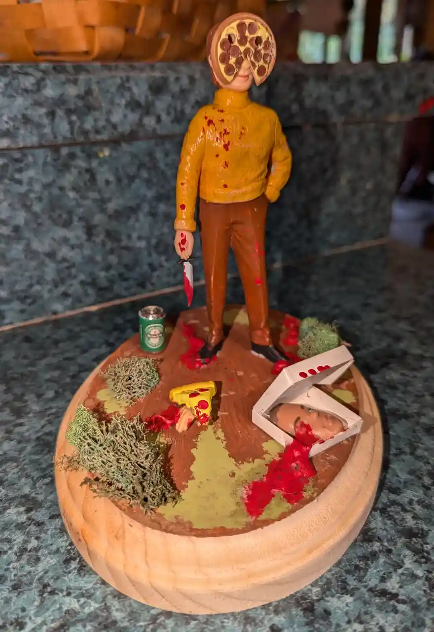 A miniature diorama depicts a figurine with a pepperoni pizza for a head, holding a knife with red paint, standing over a…