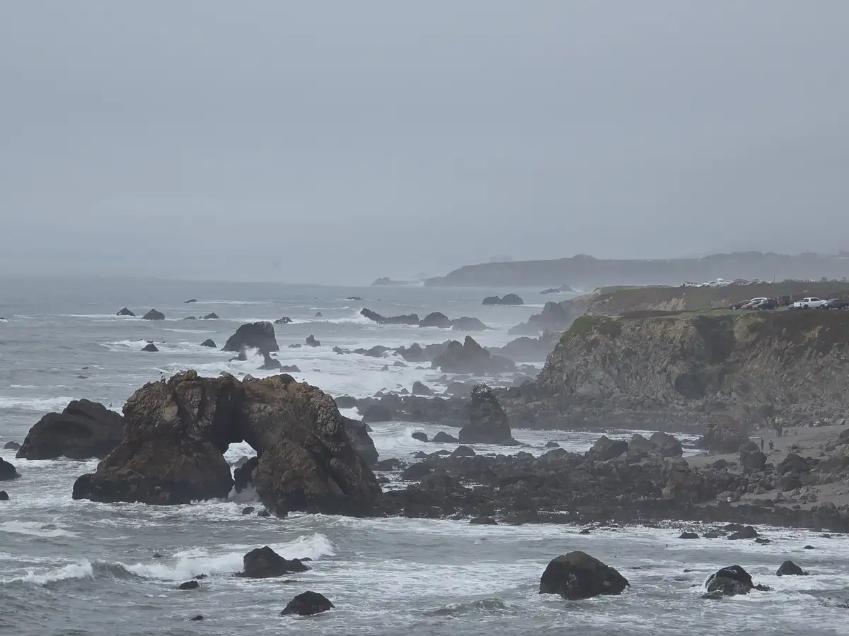 A misty coastal scene with large, dark rocks jutting from turbulent ocean waves, including a prominent rock arch. A…