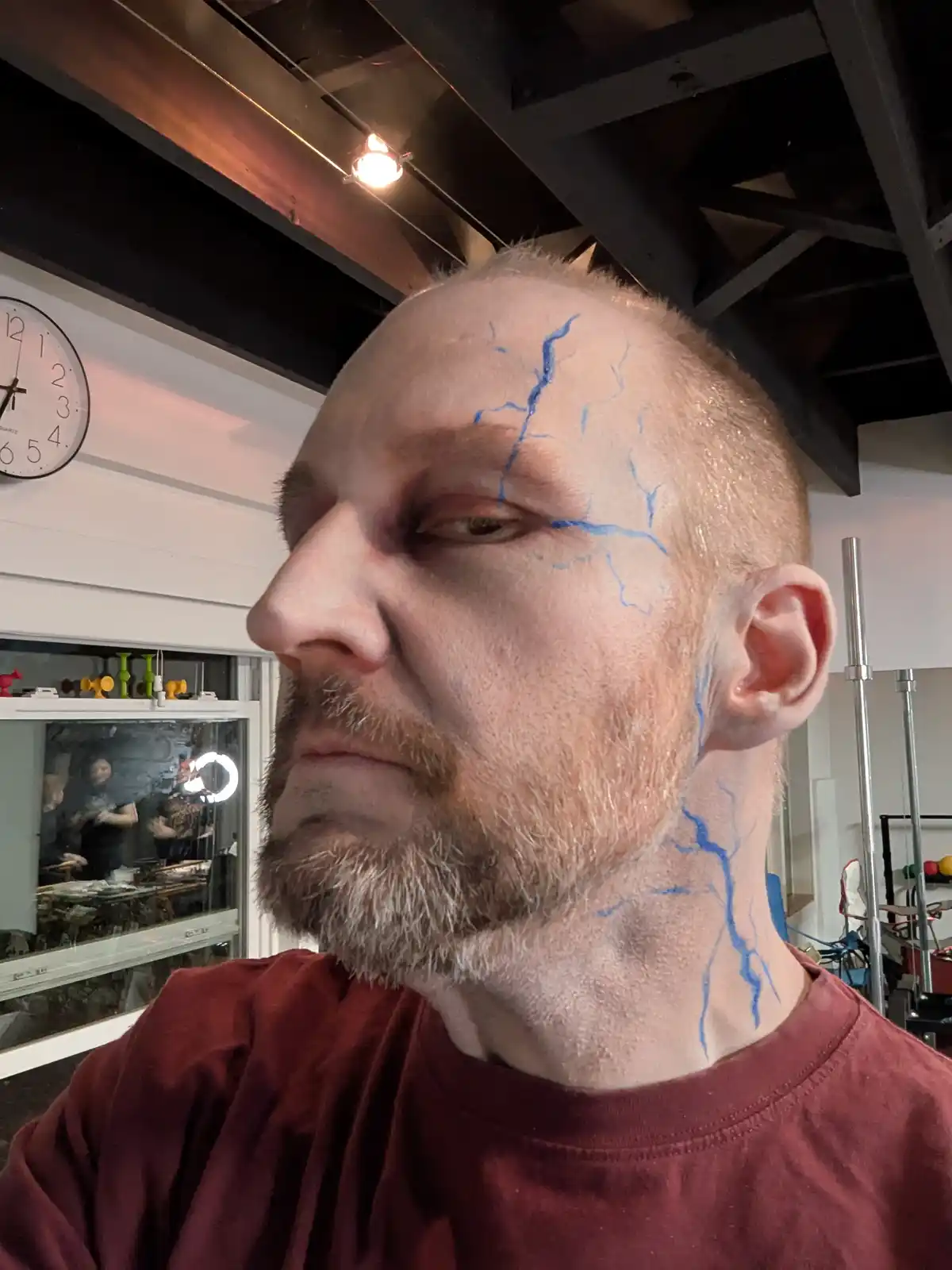 A man with a shaved head and a reddish-brown beard wears a maroon t-shirt and has blue lightning bolt drawings on his…