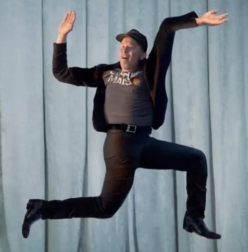 A man in a black jacket, grey t-shirt with 'Mars' text, black pants, and a baseball cap is captured mid-air in a dynamic…