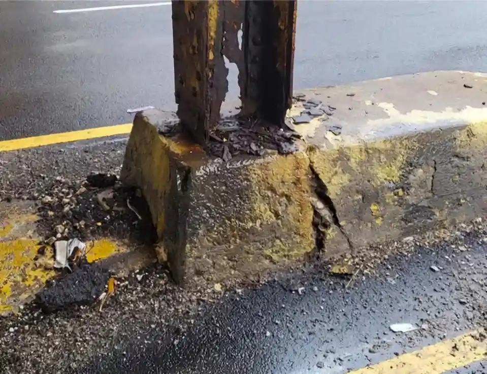 A close-up photo shows a rusty metal pole embedded in a cracked, yellow-painted concrete curb next to a wet asphalt road…