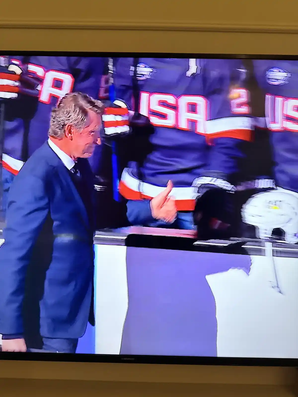 A man in a blue suit shakes hands with a hockey player in a USA jersey at an ice rink. The player's jersey has 'USA' and the…