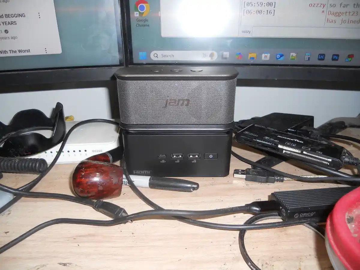 A cluttered desk with a wooden pipe, tangled cables, a router, a multi-port USB hub, and a speaker stacked on top of it…
