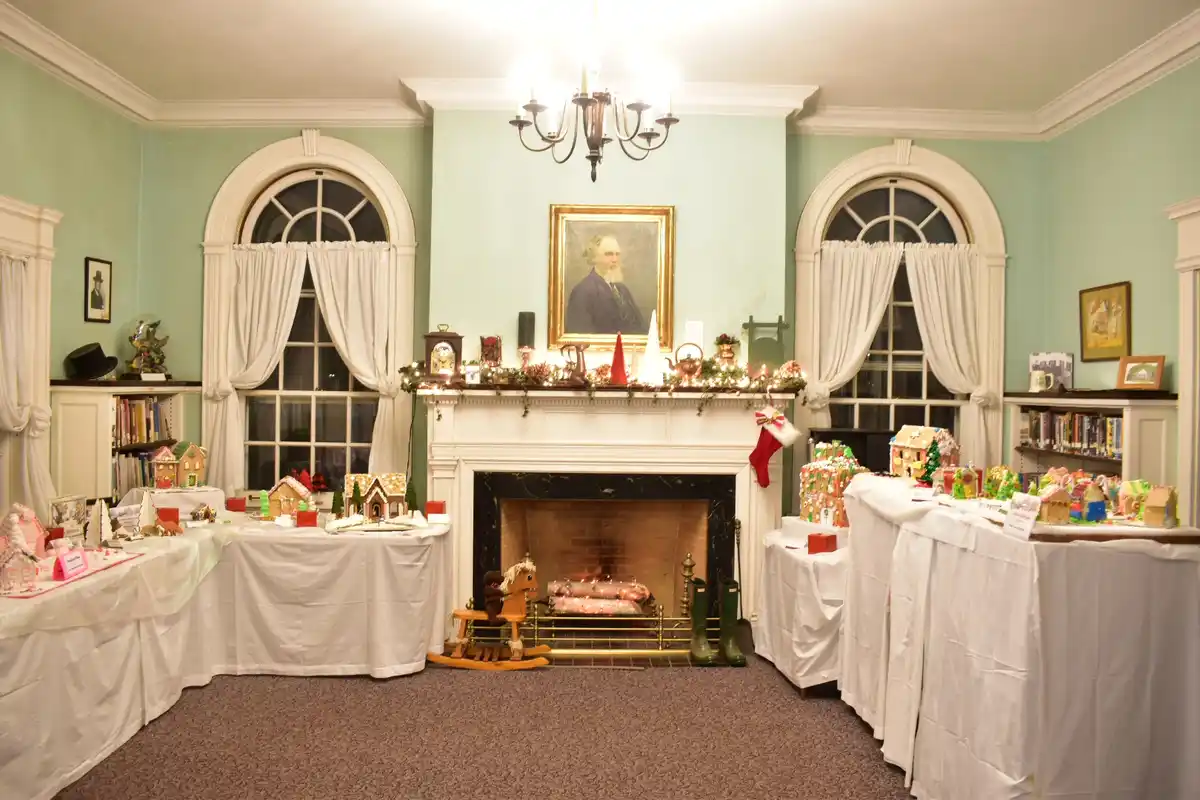 a formal room with light green walls, arched windows, and a fireplace decorated with garlands and a portrait. Several long…