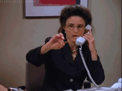 A still image from the TV show 'Seinfeld' shows a woman in a black blazer and glasses holding a white telephone receiver to…