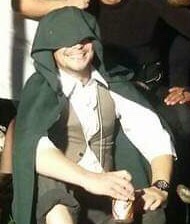 A person wearing a green hooded cloak and vest smiles while seated, holding a drink, with overlaid text reading 'i'm not a…