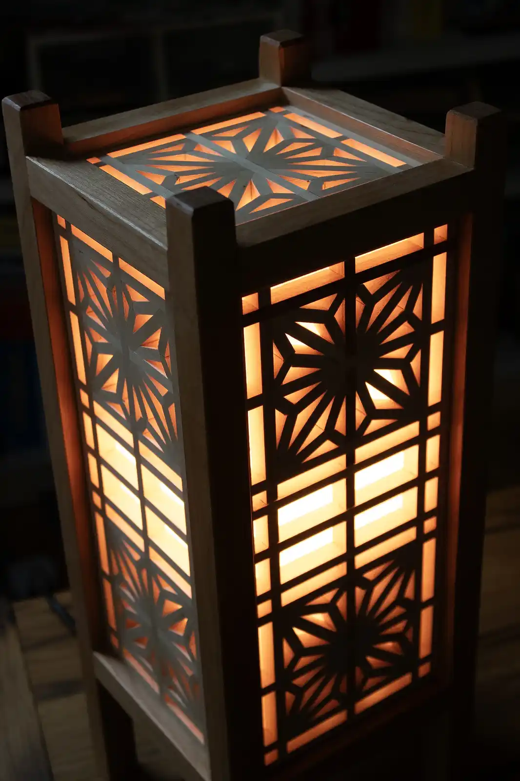 A close-up photo of a wooden, rectangular lamp with intricate geometric cutouts glowing warmly from within. The lamp has a…