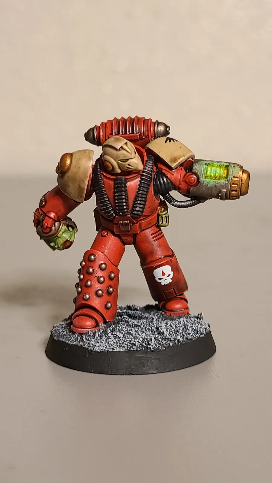 A painted miniature of a red-armored Space Marine, likely from Warhammer 40,000, stands on a textured base holding a glowing…