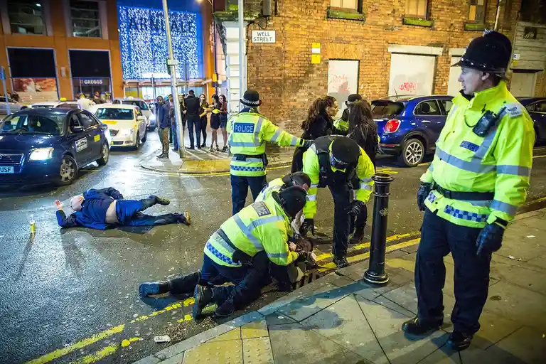 Several police officers in high-visibility jackets are attending to a man lying on the pavement in a city street at night…