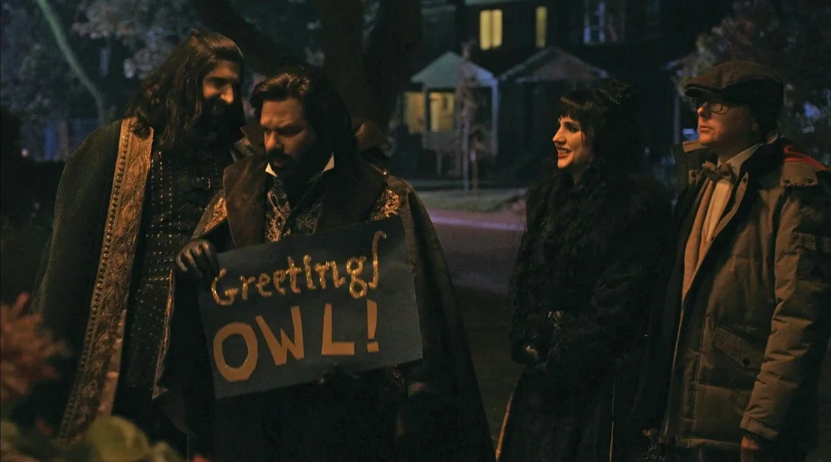 Four people stand outdoors at night, one holding a handmade sign that reads 'Greetings OWL!' in gold letters. The group…