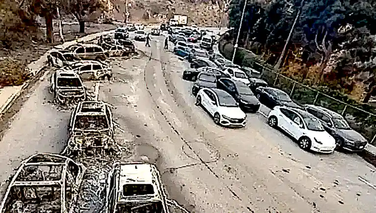 A high-angle, grainy security camera view of a steep, winding road with cars parked on both sides. The cars on the left are…