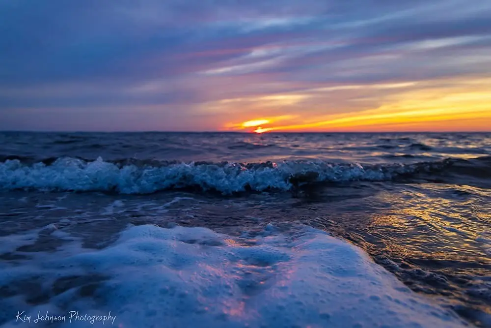 A serene sunset over the ocean with waves crashing in the foreground, displaying vibrant orange and purple hues in the sky…