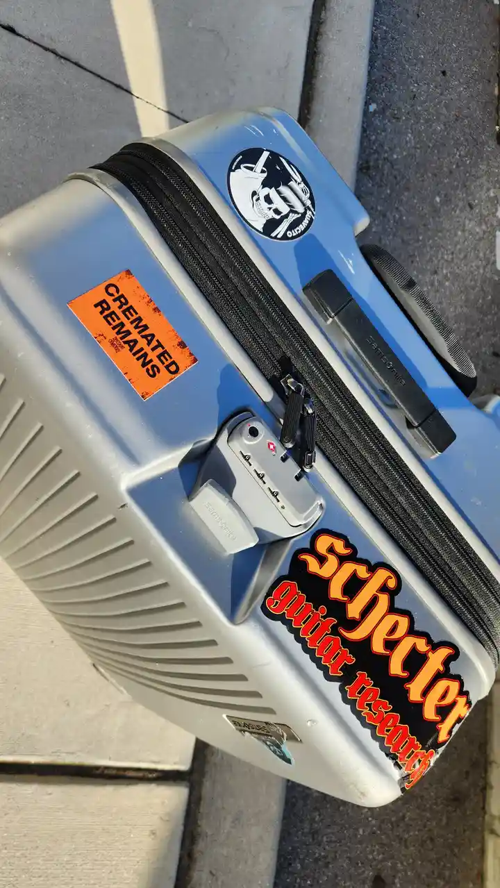 A light blue Samsonite suitcase with a 'Schechter guitar research' sticker and a skull-and-crossbones sticker sits on…
