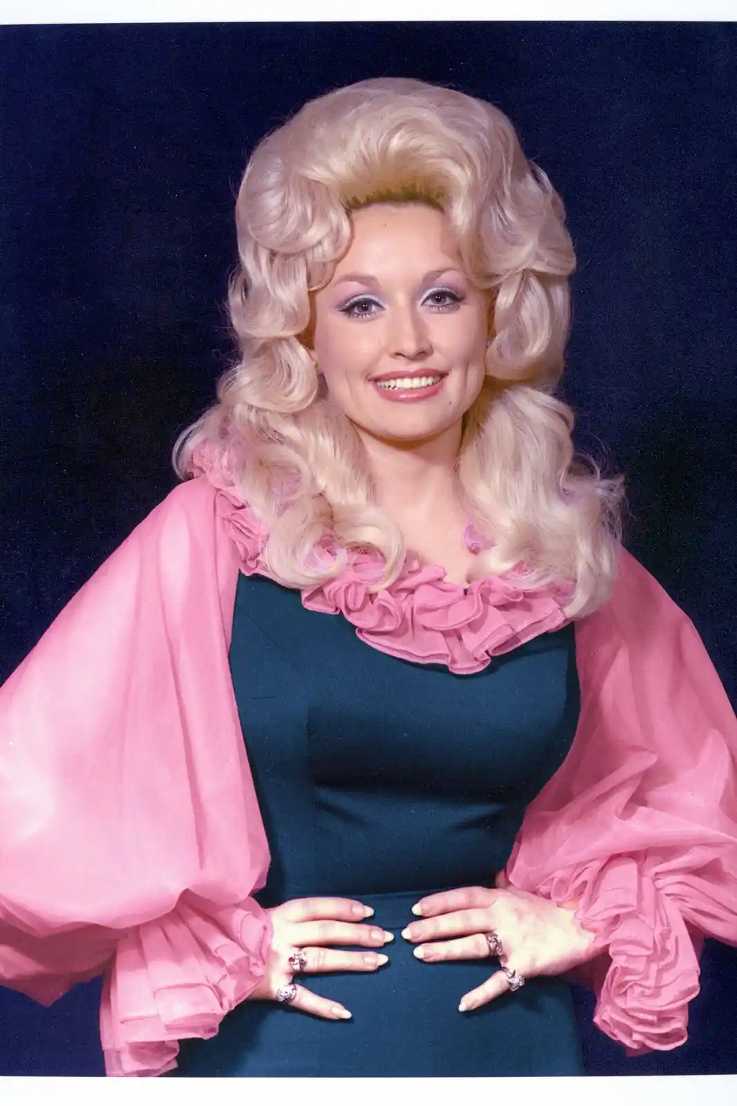 a vintage studio portrait of Dolly Parton smiling, wearing a dark teal dress with a pink ruffled collar and sheer pink bell…