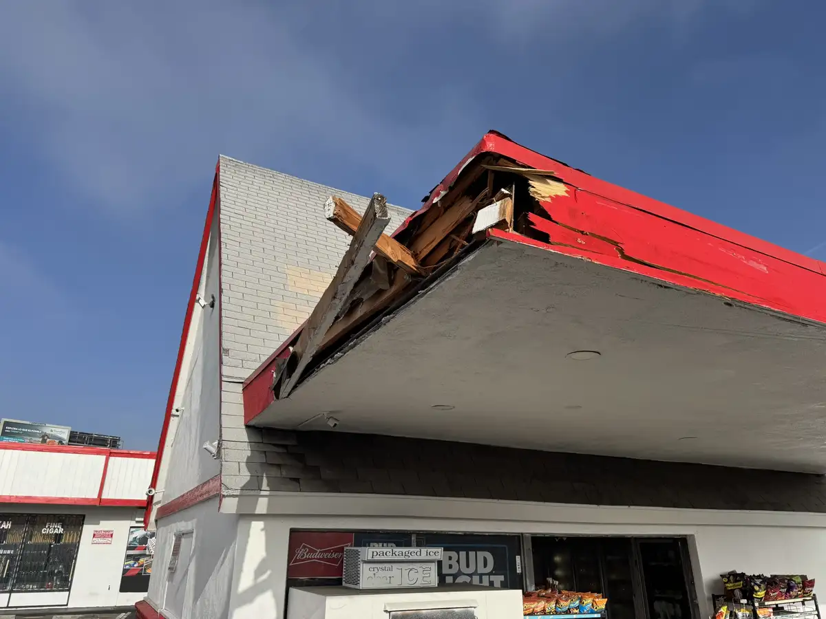 A low-angle photo shows the roof of a convenience store with red trim partially collapsed, exposing wooden beams and…